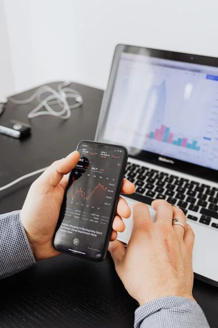 Hands using smartphone beside laptop with stock charts, showcasing digital trading.